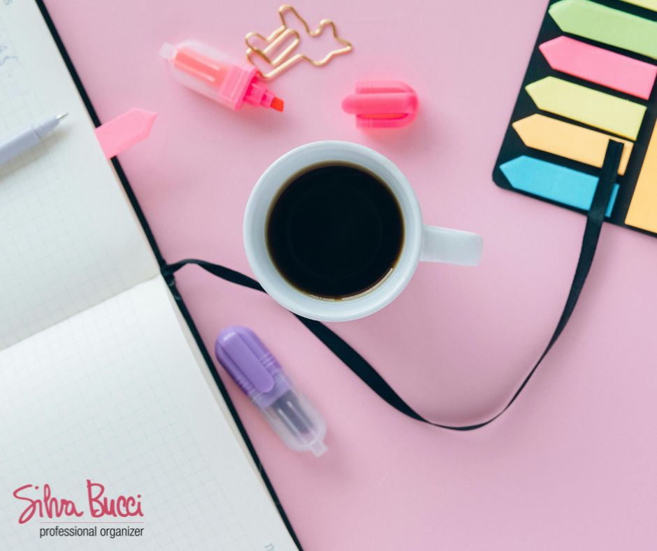 Tazza di caffè su scrivania rosa con agenda, post-it e penne colorate: un invito a fare spazio e organizzarsi prima di partire, per liberare la mente e affrontare le vacanze con leggerezza.